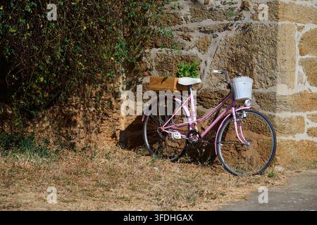 Pink vintage bicycle with a white basket and wooden crate on the rear rack leaning against an old stone wall, representing leisure, nostalgia, and sum Stock Photo