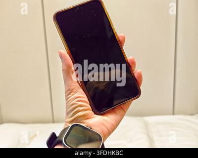 Close everyday technology scene shows a mobile phone with the display turned off held upright in one hand above a smartwatch and light sofa. Stock Photo