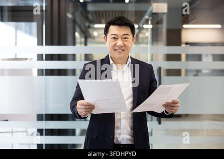 Asian businessman confidently holding business papers and smiling at the camera, standing in a modern corporate office, representing professional success, management, and corporate leadership Stock Photo