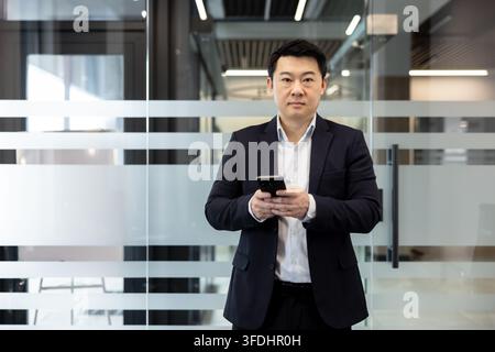Asian businessman holding a smartphone while standing in a contemporary office, representing communication, technology, professionalism, and corporate success Stock Photo