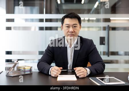 Confident asian businessman sitting at his desk, holding a pen, and looking directly at the camera, representing professionalism, leadership, and corporate success in a modern office setting Stock Photo