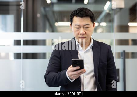 Asian businessman in a suit standing, looking at his smartphone screen, connecting with technology and managing business communication in a modern corporate office environment Stock Photo