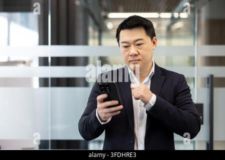 Asian businessman focused on smartphone, reading and interacting with apps in a modern office with glass partitions, representing connectivity, productivity and professional life Stock Photo