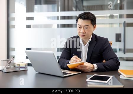 Asian businessman in a suit intently working at his office desk, looking at a laptop screen and writing notes in an orange notebook, demonstrating focus and productivity Stock Photo