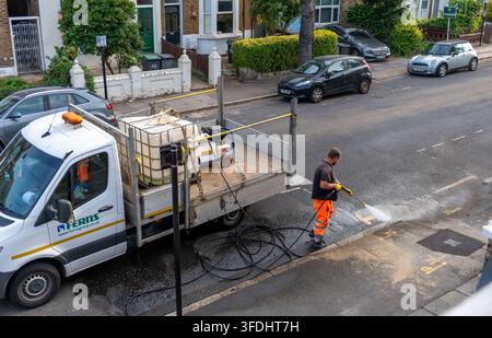 London. UK- August 28.2026. A worker using a pressure washer to clean up the mud left on the road and street after water utility work. Stock Photo