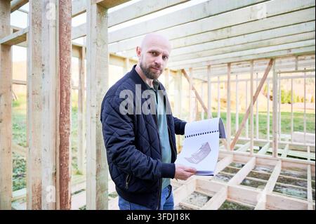 Portrait of builder shows house design drawings inside wooden frame, presenting construction concept and planning layout during early stage of residential building project Stock Photo