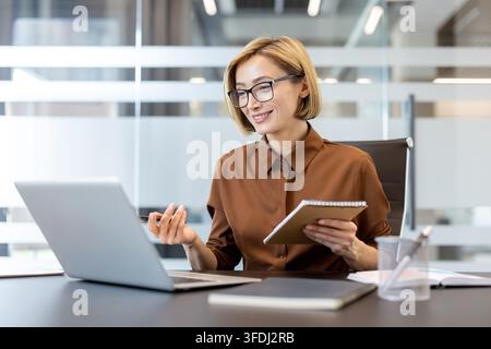 Businesswoman smiling during a video call on a laptop, holding a notebook and pen, participating in an online meeting or webinar from her modern office workspace Stock Photo