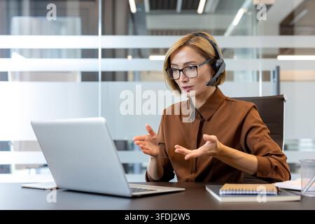 Businesswoman having a professional virtual meeting or online conference, communicating with clients or colleagues while wearing a headset and gesturing with her hands in an office environment Stock Photo