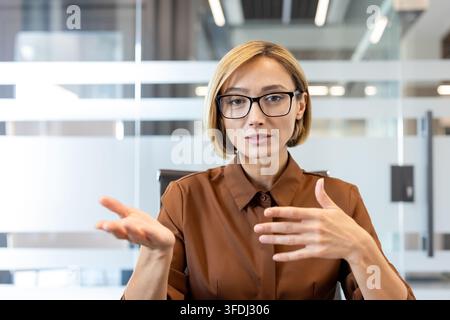 Confident businesswoman wearing glasses actively communicating and gesturing during a virtual conference or online meeting, showcasing professionalism and leadership in a modern office environment Stock Photo
