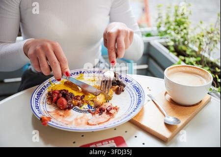 Close up of woman eating hearty breakfast at cafe, cutting sausage with eggs, beans and mushrooms, cappuccino nearby, concept of everyday lifestyle, nutrition, dining habits Stock Photo