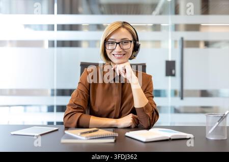 Businesswoman wearing a headset and eyeglasses, sitting at her desk, providing customer service and support, representing communication, telemarketing, and online consulting Stock Photo