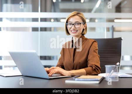 Smiling businesswoman sitting at her desk, wearing eyeglasses, using a laptop in a contemporary office environment, confidently managing tasks and representing professional success Stock Photo