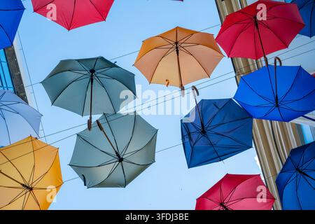 Colorful open umbrellas suspended overhead between buildings against a clear blue sky, creating a vibrant canopy of red, blue, orange, and pale gree Stock Photo