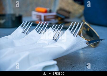 Close view of polished metal forks and a knife arranged with folded white paper napkins on a dark tabletop, with tableware in backgroun Stock Photo
