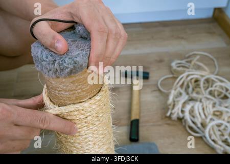 Hands repair a cat scratching post by replacing worn sisal rope around a cardboard tube, with loose cord and hand tools arranged on a wooden workbenc Stock Photo