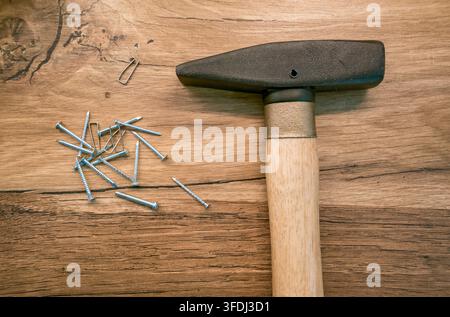 Hammer with scattered galvanized nails and a small metal fastener resting on a textured wooden workbench, viewed from above in warm ligh Stock Photo