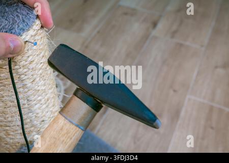 Hands secure fresh sisal rope around a worn scratching post while a hammer drives a fastener into the fiber-wrapped surface on a wooden floo Stock Photo