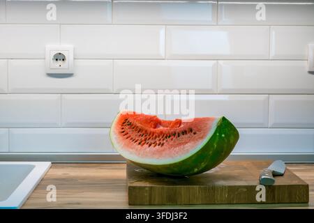 Cut watermelon half with black seeds on a wooden cutting board beside a kitchen knife in a bright kitchen with glossy white subway tile Stock Photo