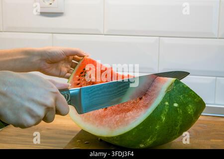 Hands slicing a fresh watermelon with a large kitchen knife on a wooden cutting board in a bright kitchen with white tiled wall Stock Photo