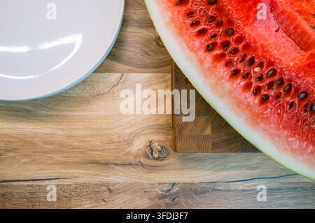 Overhead view of a seeded watermelon wedge on a wooden cutting board beside an empty white plate on a rustic wood tableto Stock Photo