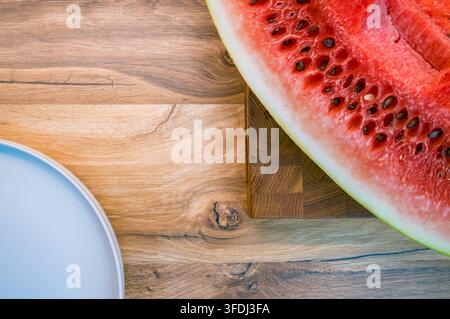 Sliced watermelon with black seeds on a wooden cutting board beside a white plate, arranged on a rustic wood countertop with open spac Stock Photo