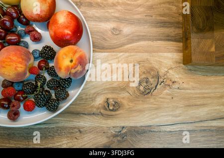 Overhead view of peaches, nectarines, raspberries, blueberries, cherries, and blackberries arranged on a white plate beside a wooden cutting boar Stock Photo