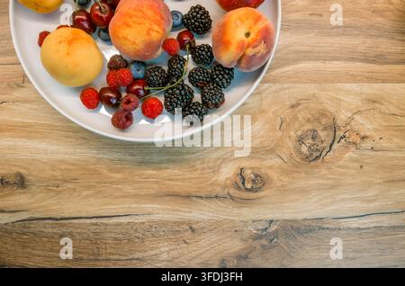 Top view of peaches, nectarines, raspberries, blueberries, cherries, and blackberries arranged on a white plate over a wooden counterto Stock Photo