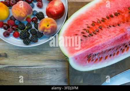 Top view of sliced watermelon on a wooden board beside a plate of peaches, nectarines, raspberries, blueberries, cherries, and blackberries Stock Photo