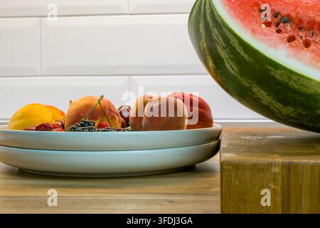 Fresh watermelon half on a wooden cutting board beside a white plate of peaches, nectarines, raspberries, blueberries, cherries, and blackberrie Stock Photo