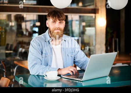 Happy remote worker using laptop in spacious loft style cafe, direct gaze and calm posture illustrating digital profession, flexible schedule, and modern workplace concept Stock Photo