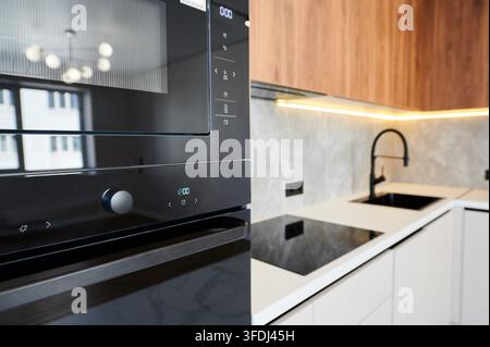 Built in oven and microwave beside cooktop and sink in modern kitchen interior. Minimal white cabinetry with wooden accents, LED lighting and premium appliance design concept Stock Photo
