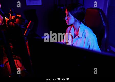 Woman focusing on her computer screen, surrounded by neon red and blue lighting, representing cybersecurity, coding, or gaming Stock Photo