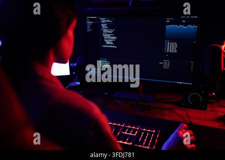 Hacker working on computer in dark room, writing code for cybersecurity attack or data breach, showing data on screen Stock Photo
