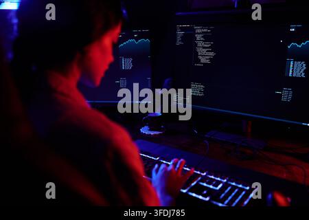 Hacker working late at night, typing on a glowing keyboard with multiple screens displaying code and data Stock Photo