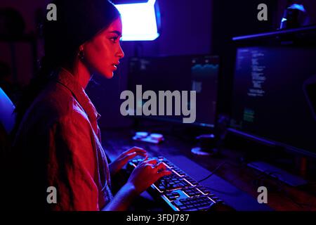 Young woman working on computer, typing code on keyboard in a dark room with vibrant red and blue neon lights Stock Photo