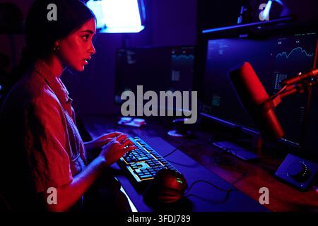 Woman programmer working on computer with multiple monitors, typing on keyboard in dark room with cyber neon light Stock Photo