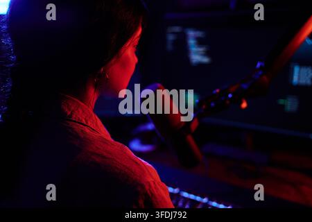 Female programmer focused on screen, working with computer code and microphone, surrounded by neon red and blue light Stock Photo