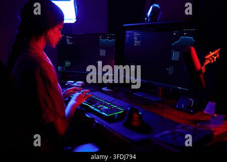 Woman programmer working on multiple computer screens with data and code, focusing on cybersecurity in a dark environment Stock Photo