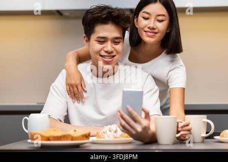 Cheerful Vietnamese Couple Using Smartphone During Breakfast In Modern Kitchen Stock Photo