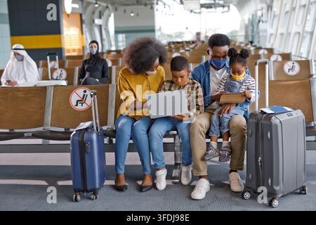 A Black family with two children sits in an airport terminal, wearing face masks and using electronic devices while waiting for their flight Stock Photo