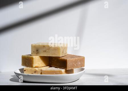 Stack of handmade artisan soap bars arranged on a white ceramic plate in soft natural light. The rustic bars feature warm neutral tones and natural va Stock Photo