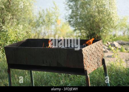 Outdoor barbecue grill with bright fire flames from burning charcoal on a sandy riverbank with green trees background in summer. Stock Photo