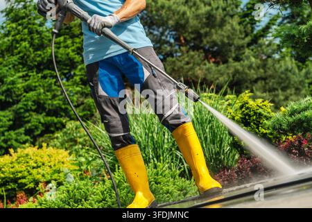 A worker in gloves and rubber boots holds a pressure washer lance at an angle. A concentrated stream of water cleans paving beside a landscaped garden Stock Photo