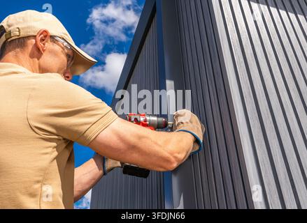 A worker wearing safety glasses and gloves drills into an exterior wall panel. The close side view highlights the tool, hands and vertical cladding. Stock Photo