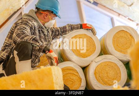 A worker wearing a dust mask, safety glasses and a hard hat handles stacked insulation rolls. Insulation fitted between the rafters fills the surround Stock Photo