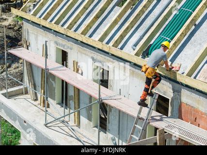 A worker in a hard hat stands on a ladder near the eaves of a house under construction. Scaffolding, timber roof members and bundled pipes are visible Stock Photo