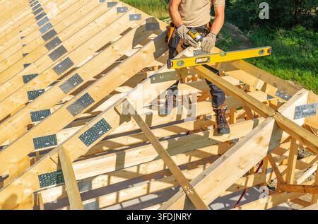 An elevated close view shows a carpenter using a cordless driver on a metal truss connector. A spirit level rests across the surrounding timber roof m Stock Photo
