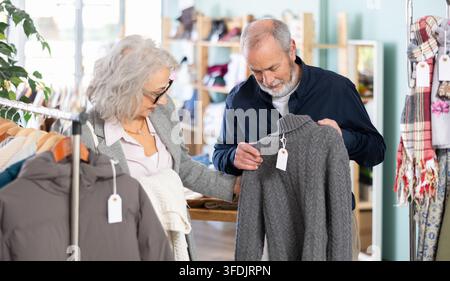 Mature man and woman standing in a clothing store and choosing knitted sweaters together. Stylish pensioners go shopping in the mall Stock Photo
