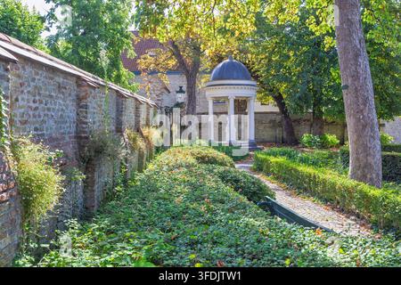 Bruges town in Belgium, Europe. Charming garden path winds past a historic stone wall, lush greenery, and a delightful classical gazebo under bright Stock Photo