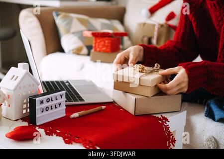 Hands stack kraft boxes beside laptop on table with HOLLY JOLLY sign and ceramic house. Returning unwanted christmas gift, gift exchange policy, post Stock Photo
